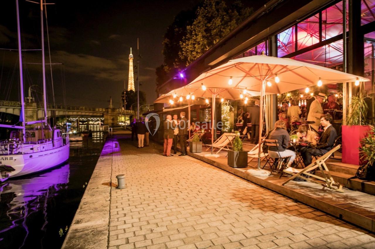 Location salle Paris 15 (Paris) - Paris Yacht Marina #21 Terrasse animée d'un restaurant au bord de l'eau, éclairée par des guirlandes lumineuses, avec la Tour Eiffel en arrière…