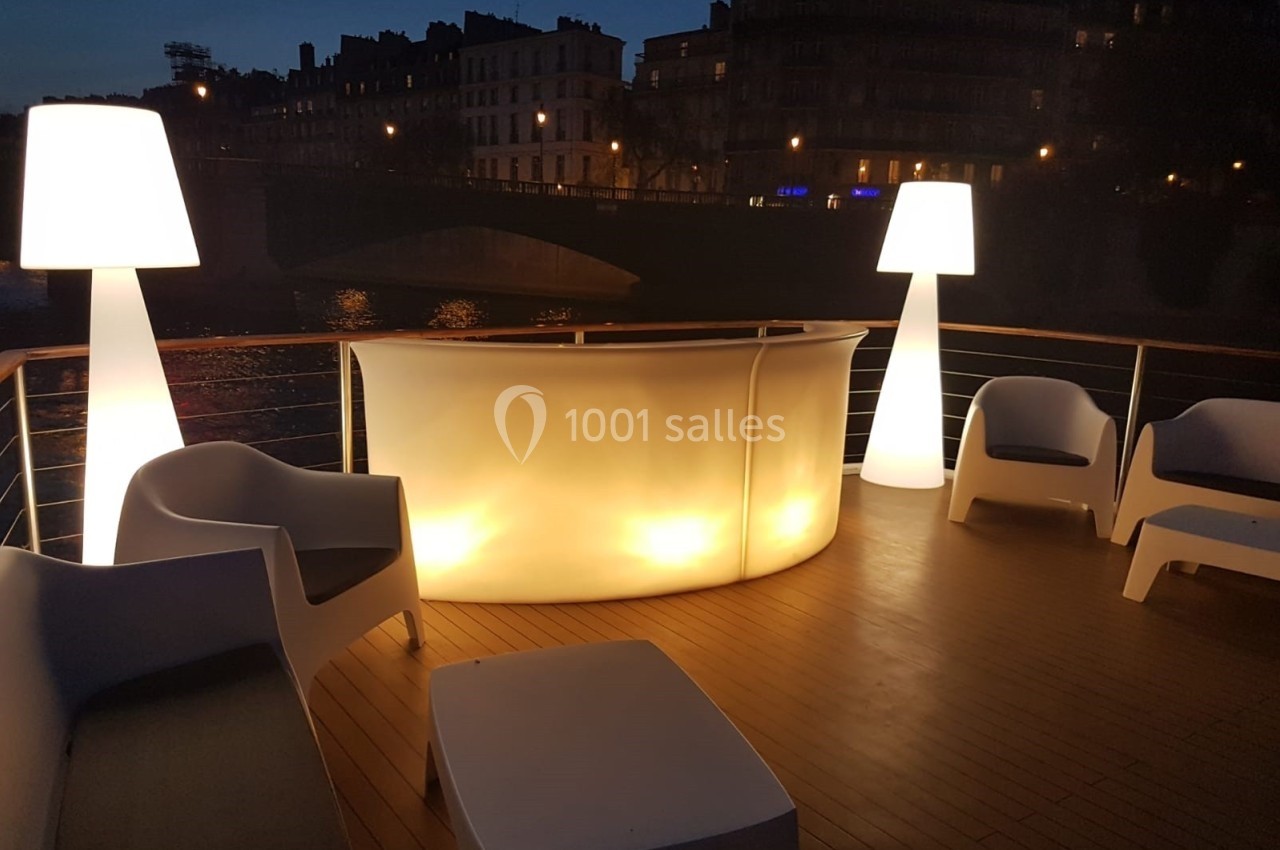 Location salle Paris 15 (Paris) - Paris Yacht Marina #28 Terrasse en soirée avec un bar lumineux, fauteuils blancs et lampes design, vue sur des bâtiments et un pont éclairés.