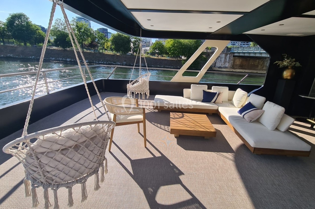Location salle Paris 15 (Paris) - Paris Yacht Marina #38 Espace extérieur d'un bateau avec fauteuils suspendus, canapé blanc, table basse en bois et vue sur une rivière bordée d…