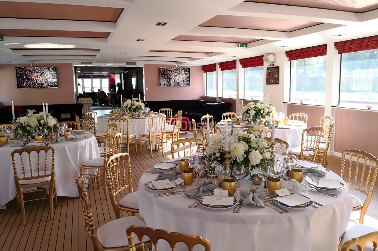 Salle de réception élégamment décorée avec des tables rondes, nappes blanches, chaises dorées et arrangements floraux.