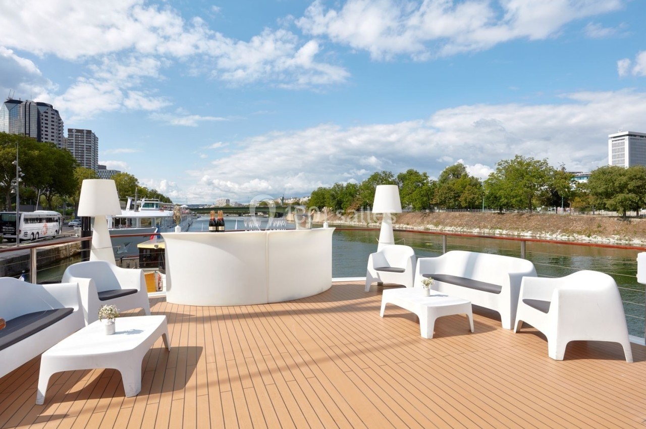 Terrasse d'un bateau avec mobilier blanc moderne, vue sur un fleuve bordé d'arbres et de bâtiments urbains.