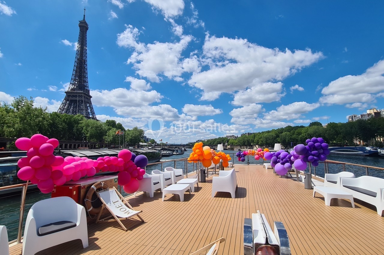 Pont d'un bateau sur la Seine décoré de ballons colorés, avec vue sur la tour Eiffel par temps ensoleillé.