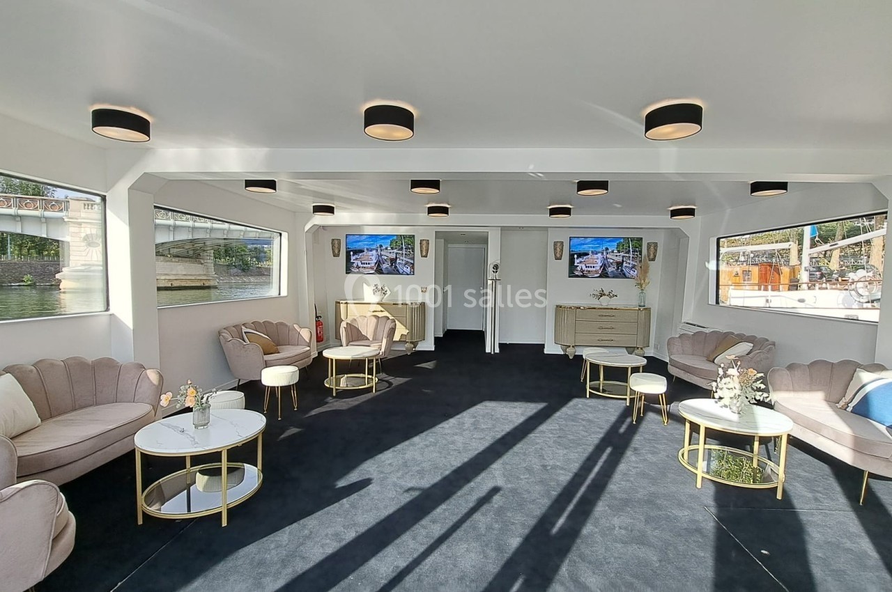 Salon lumineux avec canapés beiges, tables basses dorées et grandes fenêtres offrant une vue sur l'extérieur.