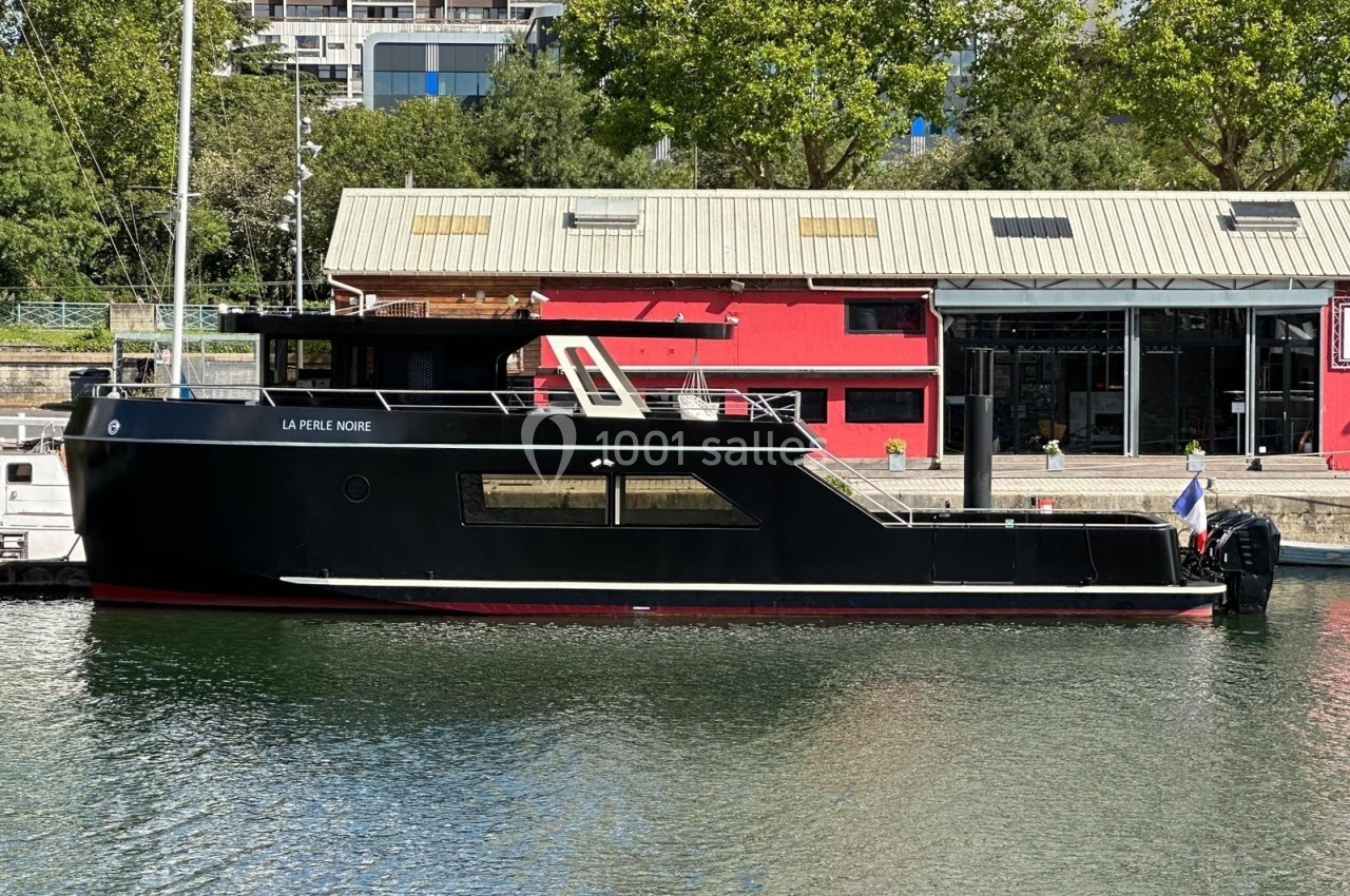 Bateau noir amarré près d'un quai avec un bâtiment rouge et des arbres en arrière-plan.