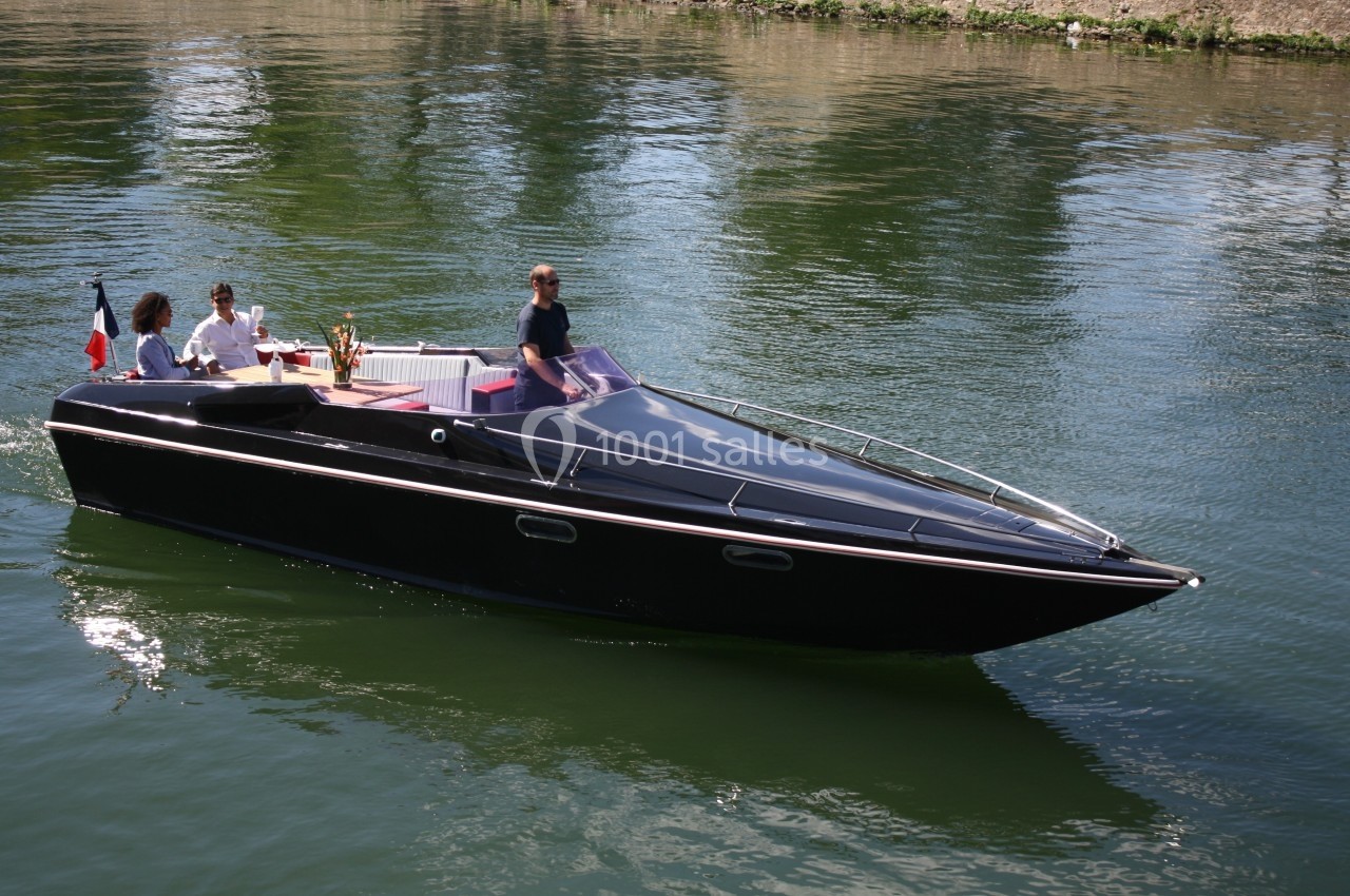 Un bateau à moteur noir navigue sur une rivière avec trois personnes à bord, dont une tenant une bouteille.