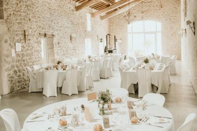 Salle de réception lumineuse avec tables rondes dressées, nappes blanches, chaises en bois et décorations florales.