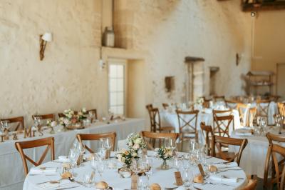 Salle de réception lumineuse avec tables rondes dressées, nappes blanches, chaises en bois et décorations florales.