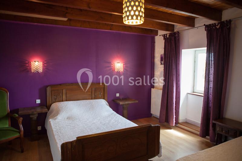 Chambre avec murs violets, lit en bois, couvre-lit blanc, fauteuil vert, poutres apparentes et lumière naturelle.