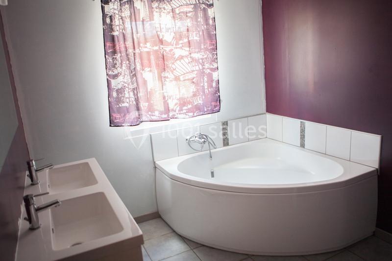 Salle de bain avec baignoire d'angle blanche, double vasque et mur violet, éclairée par une fenêtre avec rideau.