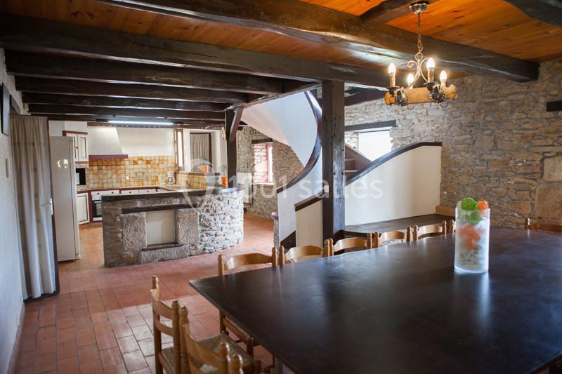Salle à manger rustique avec table en bois, chaises assorties, cuisine ouverte en pierre et poutres apparentes au plafond.