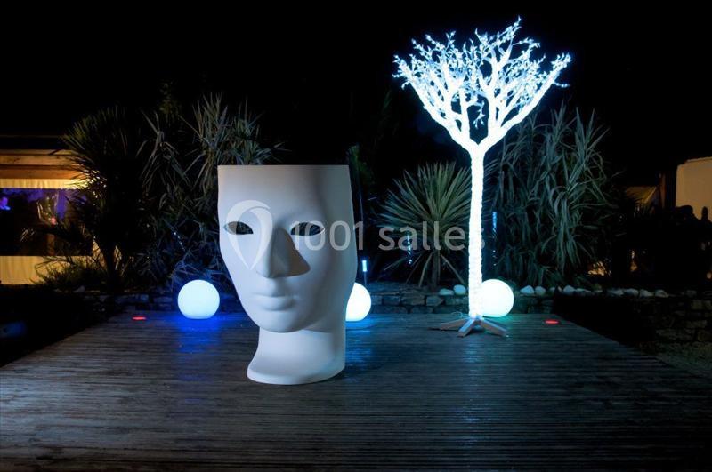 Sculpture en forme de visage blanc devant un arbre lumineux et des sphères éclairées, dans un décor nocturne.