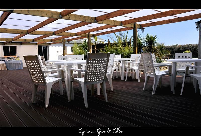 Terrasse en bois avec tables et chaises blanches sous une pergola transparente, entourée de végétation.