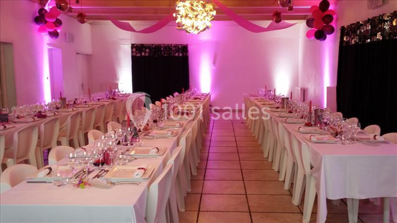 Salle décorée pour un événement avec deux longues tables dressées, éclairage rose et guirlandes suspendues.