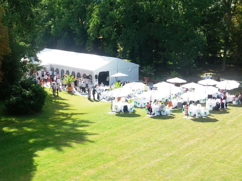 Vue aérienne d'un événement en plein air avec des tables sous des parasols et une grande tente blanche dans un parc…