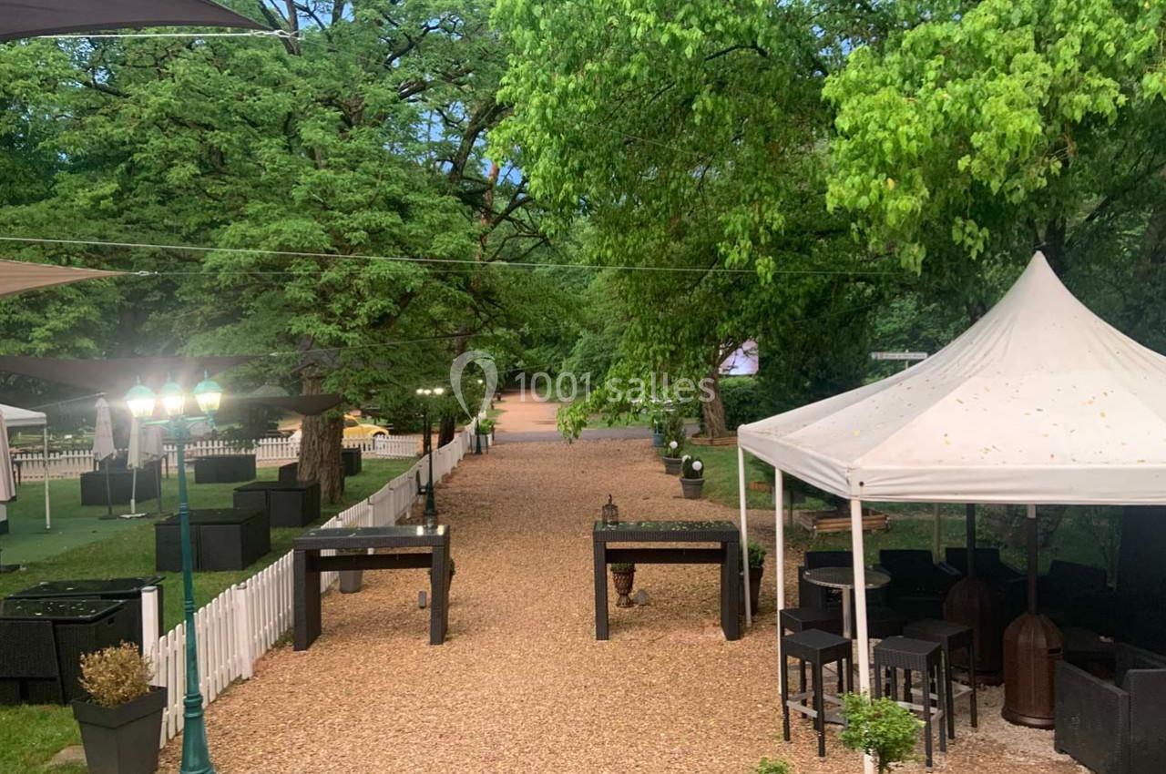 Allée en gravier bordée de tables, chaises et parasols, entourée de verdure et clôtures blanches.