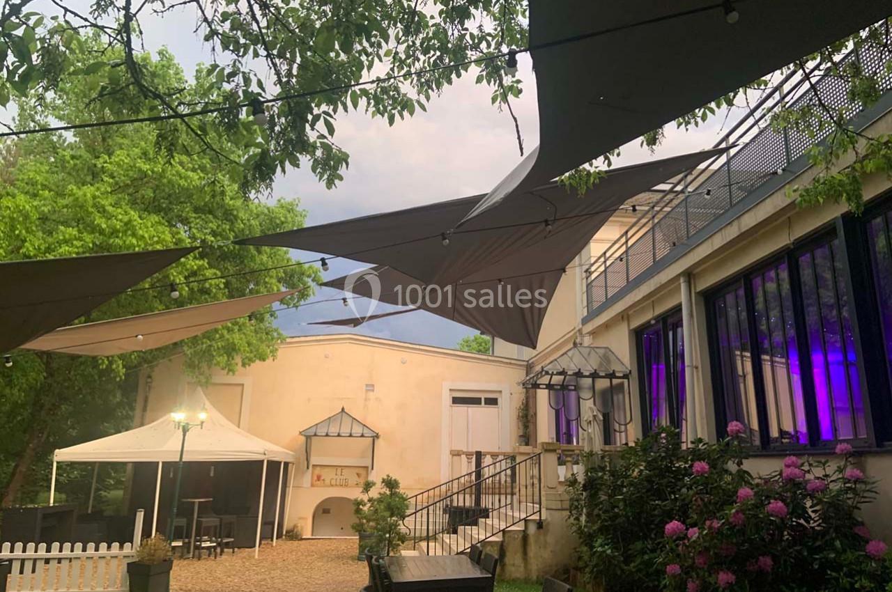 Cour extérieure avec voiles d'ombrage, mobilier de jardin, plantes fleuries et éclairage tamisé près d'un bâtiment clair.