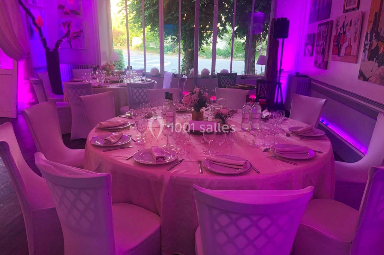 Salle décorée pour un événement avec tables rondes dressées, nappes roses, chaises blanches et éclairage violet.