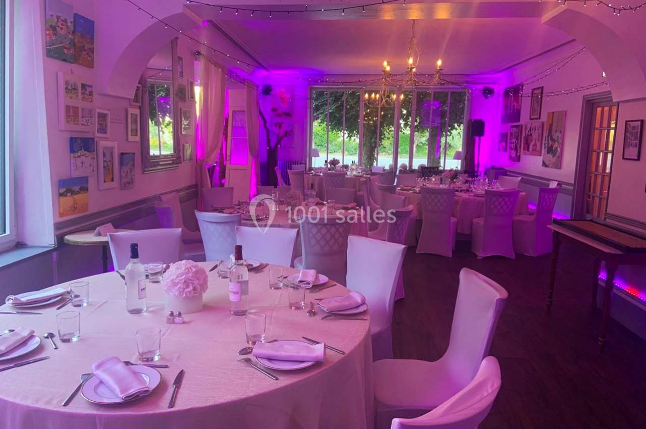 Salle de réception décorée avec des tables rondes dressées, éclairage rose et vue sur un jardin à travers de grandes…
