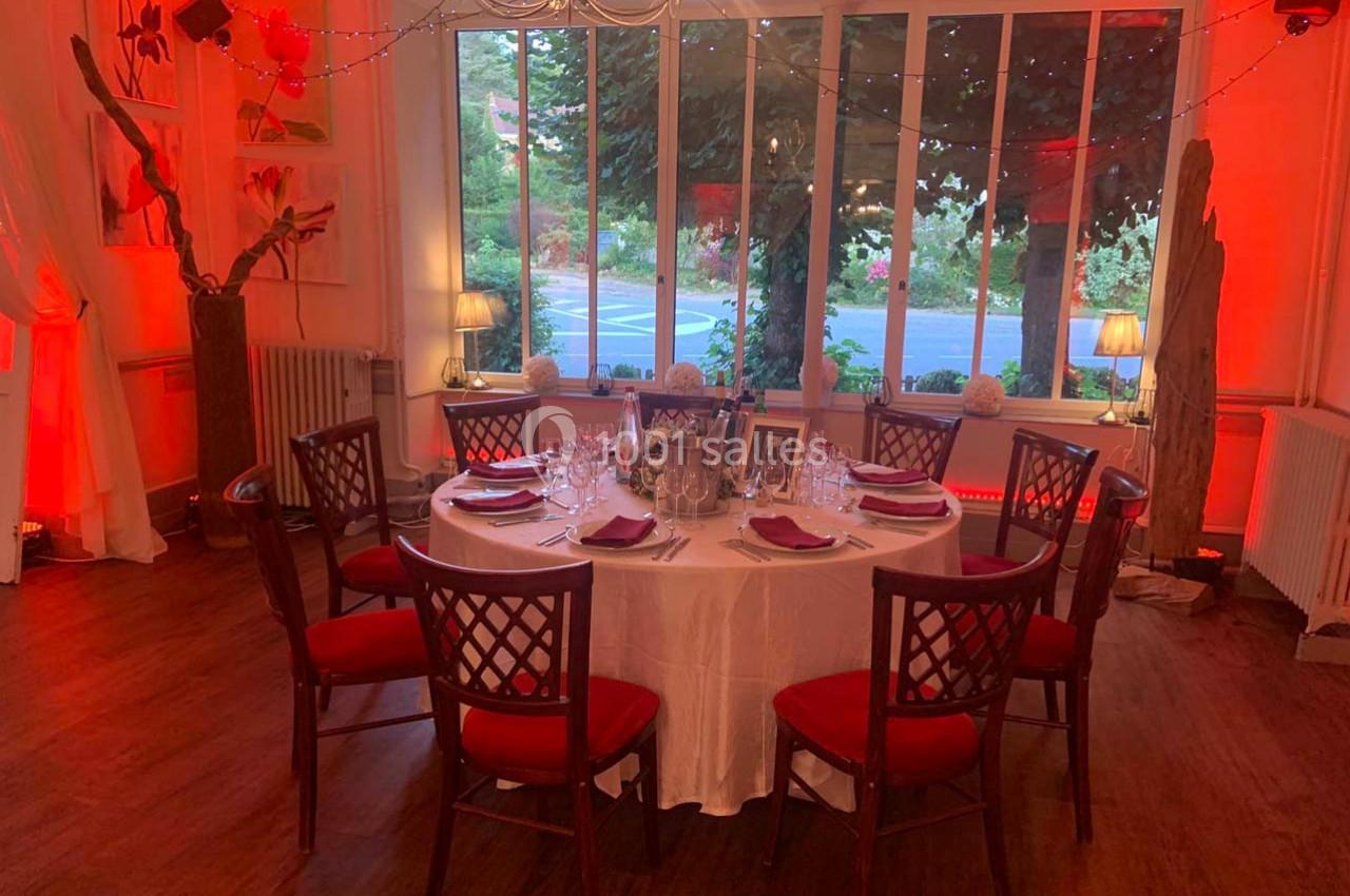 Salle décorée avec une table ronde dressée pour un repas, entourée de chaises en bois avec coussins rouges.