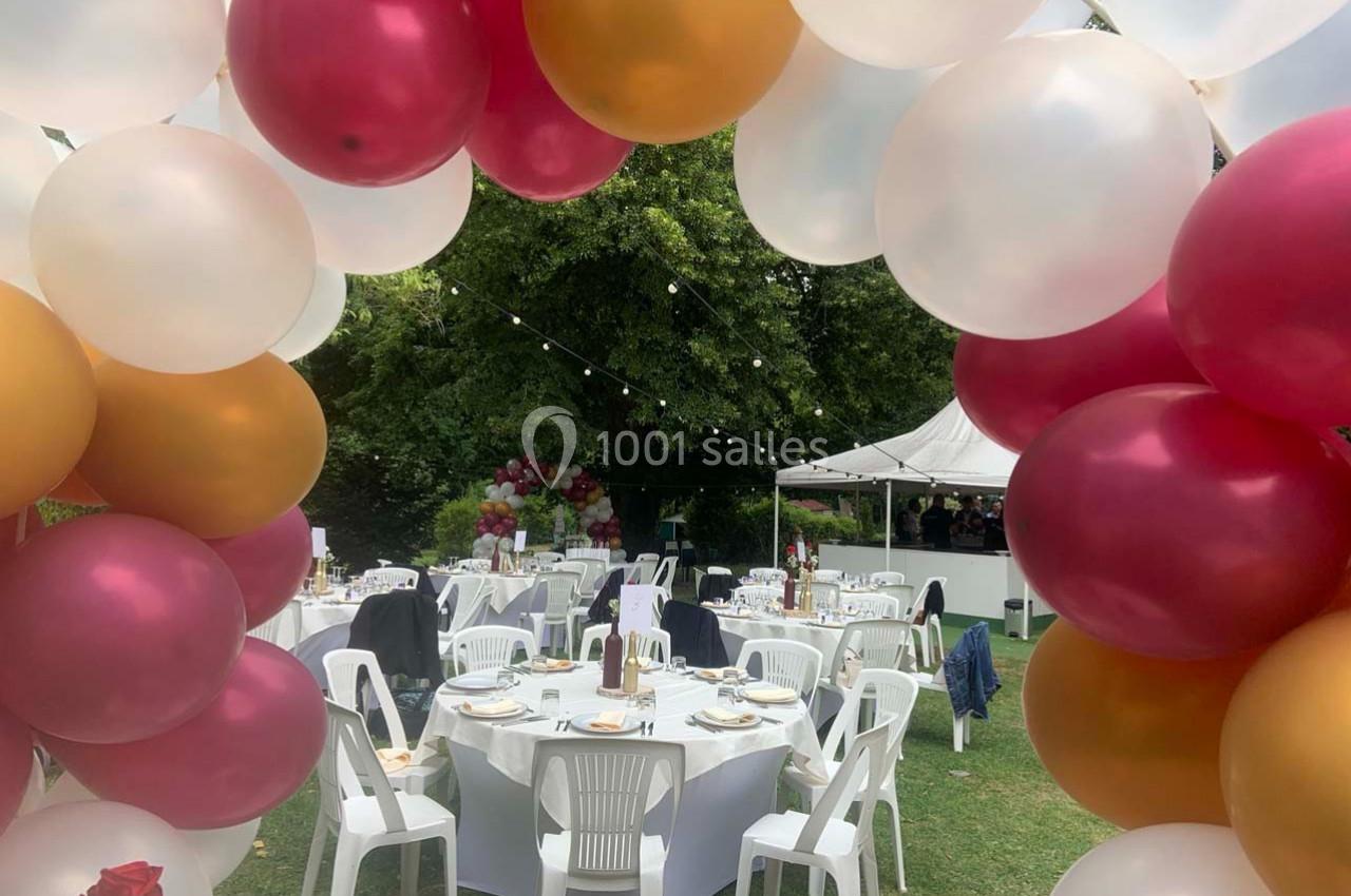 Arche de ballons colorés encadrant des tables dressées pour un événement en plein air dans un jardin.