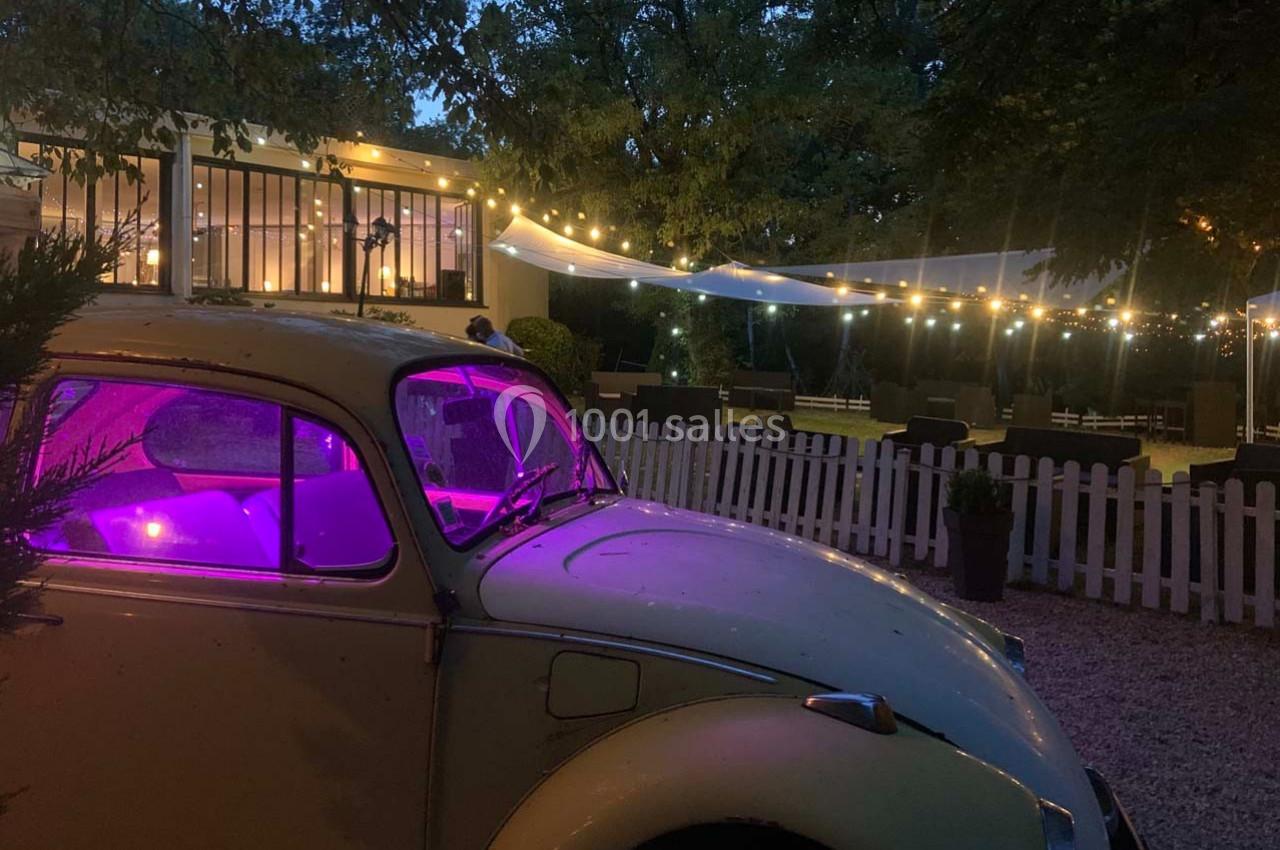 Voiture ancienne éclairée en violet devant une terrasse illuminée et décorée de guirlandes lumineuses, en soirée.