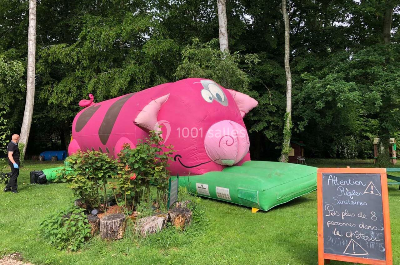 Structure gonflable rose en forme de cochon dans un parc boisé, avec un panneau indiquant des consignes de sécurité.