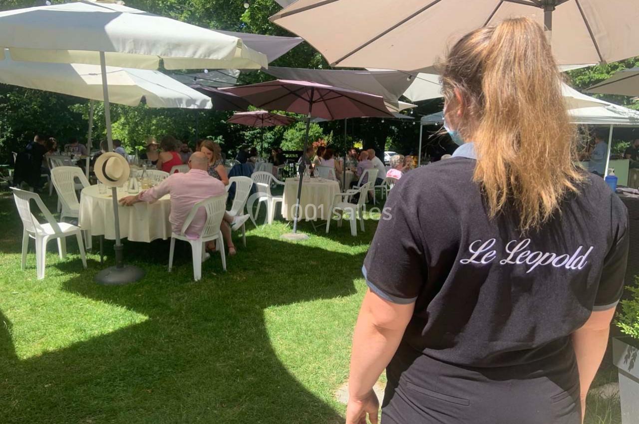 Serveuse observant des clients attablés en terrasse ombragée, entourée de parasols et de verdure.