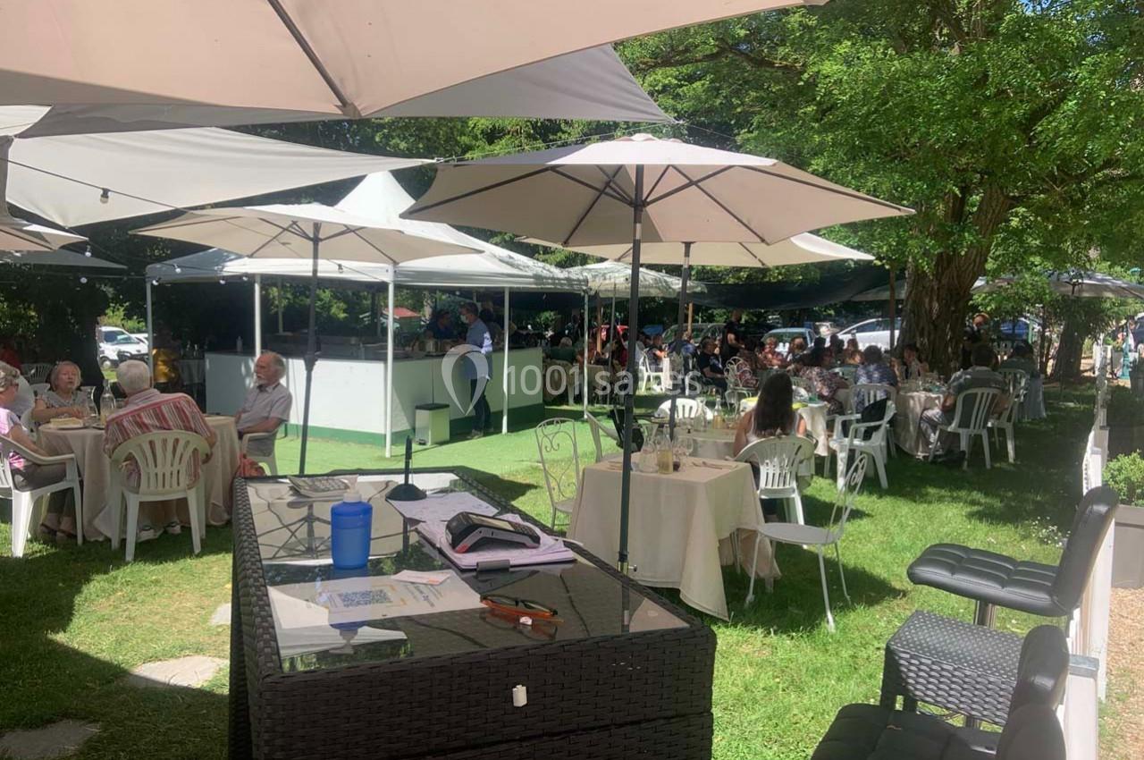 Des personnes déjeunent en plein air sous des parasols dans un espace verdoyant avec des tables et un bar en arrière-plan.