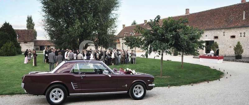 Voiture ancienne rouge garée devant un espace vert où des invités se rassemblent près d'un bâtiment en pierre.