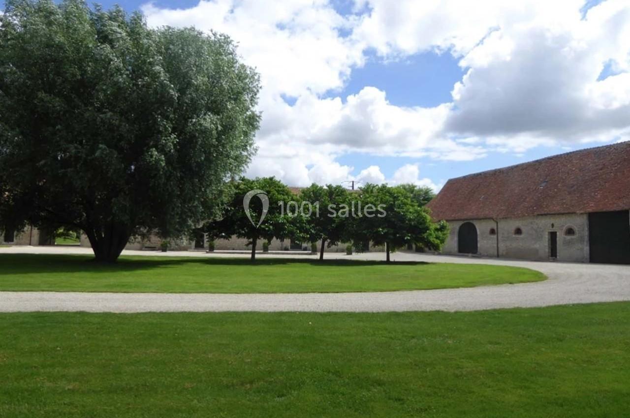 Cour herbeuse avec un chemin gravillonné, entourée d'arbres et de bâtiments en pierre sous un ciel partiellement nuageux.