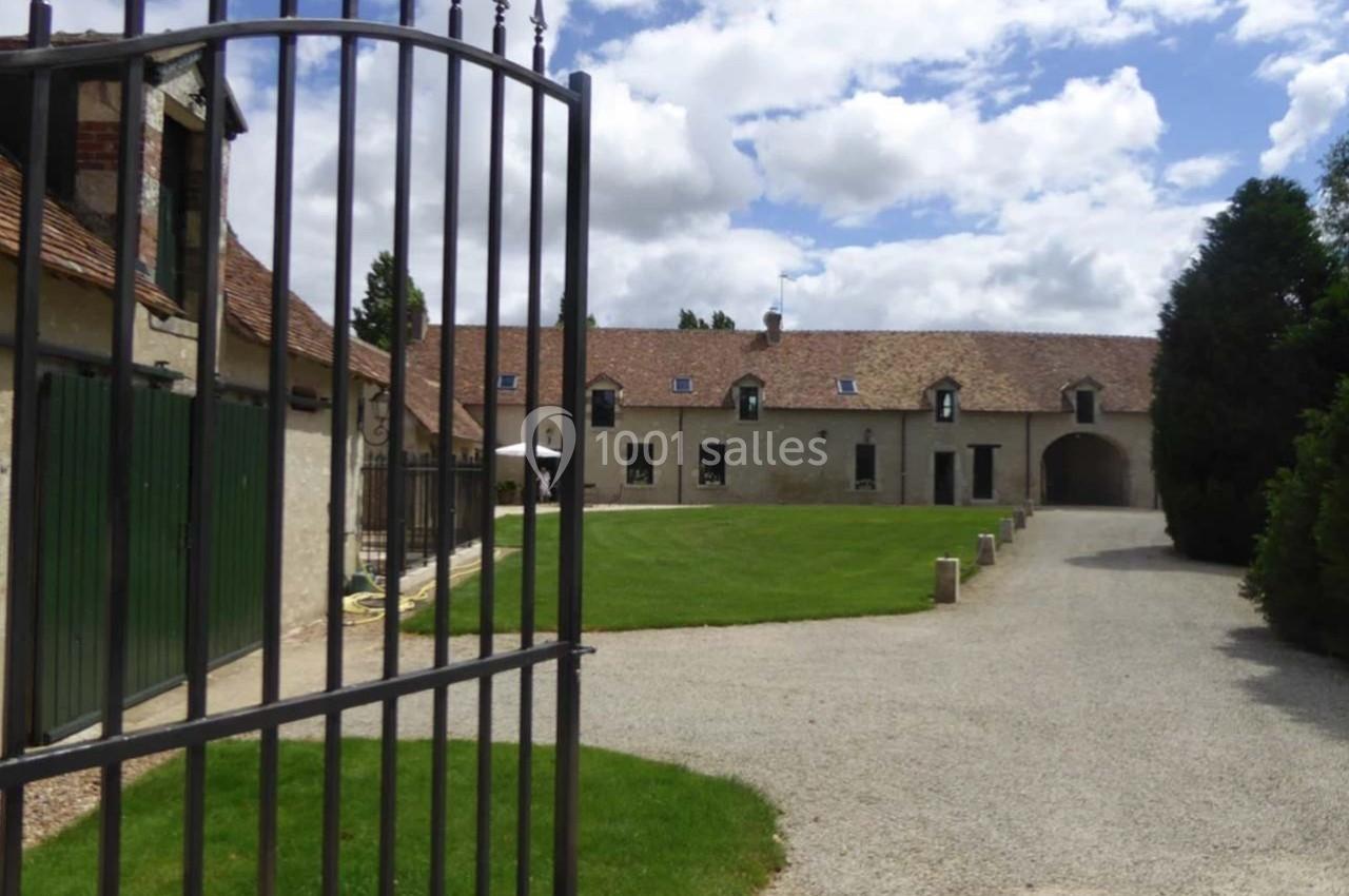 Cour d'une grande bâtisse en pierre avec toit en tuiles, vue depuis un portail en fer forgé, ciel partiellement nuageux.