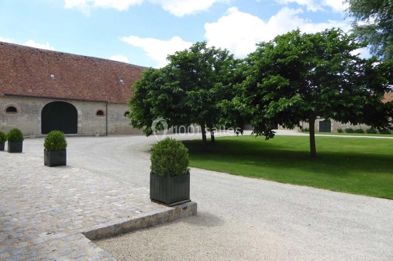 Cour pavée avec arbres et pelouse, bordée de bâtiments en pierre aux toits en tuiles rouges sous un ciel dégagé.