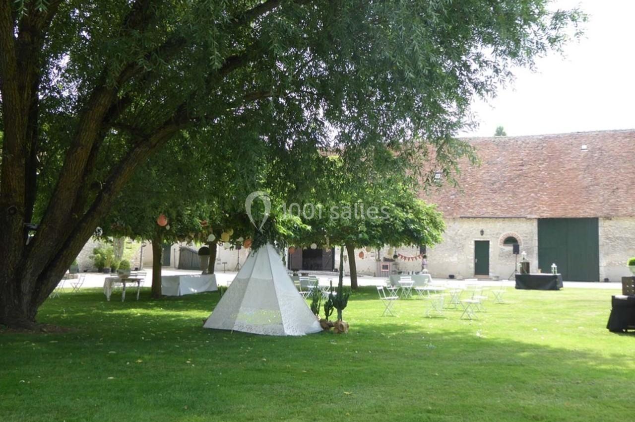 Jardin avec pelouse, grand arbre, tipi blanc, tables et chaises, devant des bâtiments en pierre et une grange.