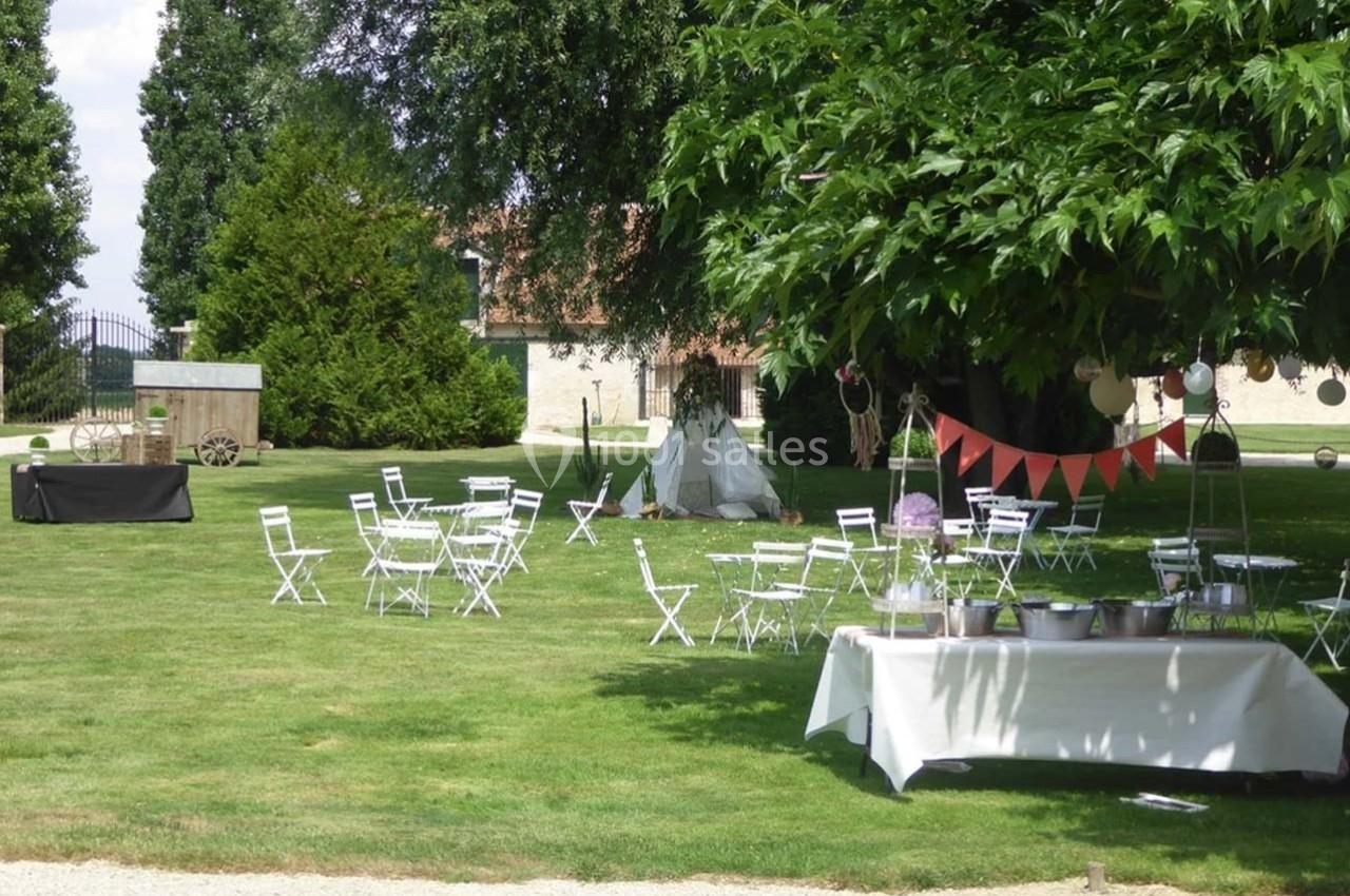 Jardin avec des tables et chaises blanches disposées pour un événement, décorations et tente tipi en arrière-plan.