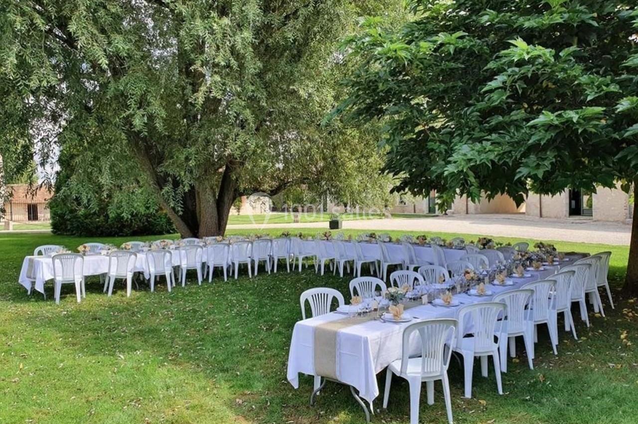 Tables dressées avec nappes blanches et chaises en plastique disposées sous des arbres dans un jardin verdoyant.