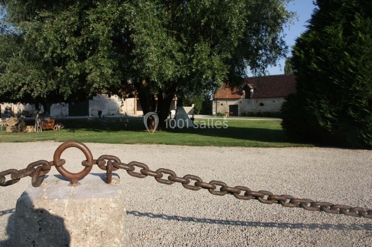 Chaîne en métal attachée à un poteau en pierre, avec en arrière-plan un jardin, des arbres et des bâtiments anciens.