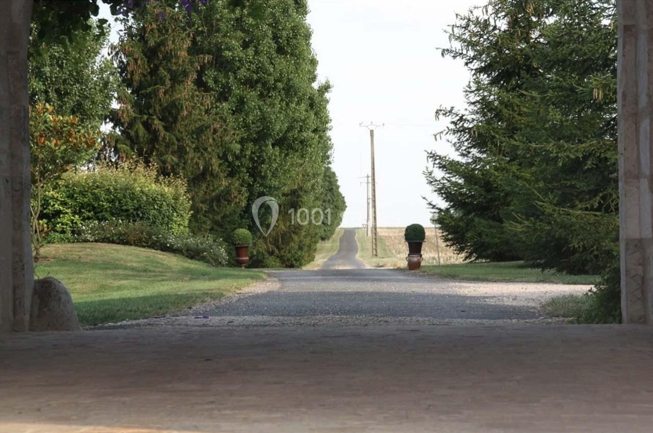 Allée bordée d'arbres et de pelouses, vue depuis un porche, menant à une route de campagne sous un ciel dégagé.