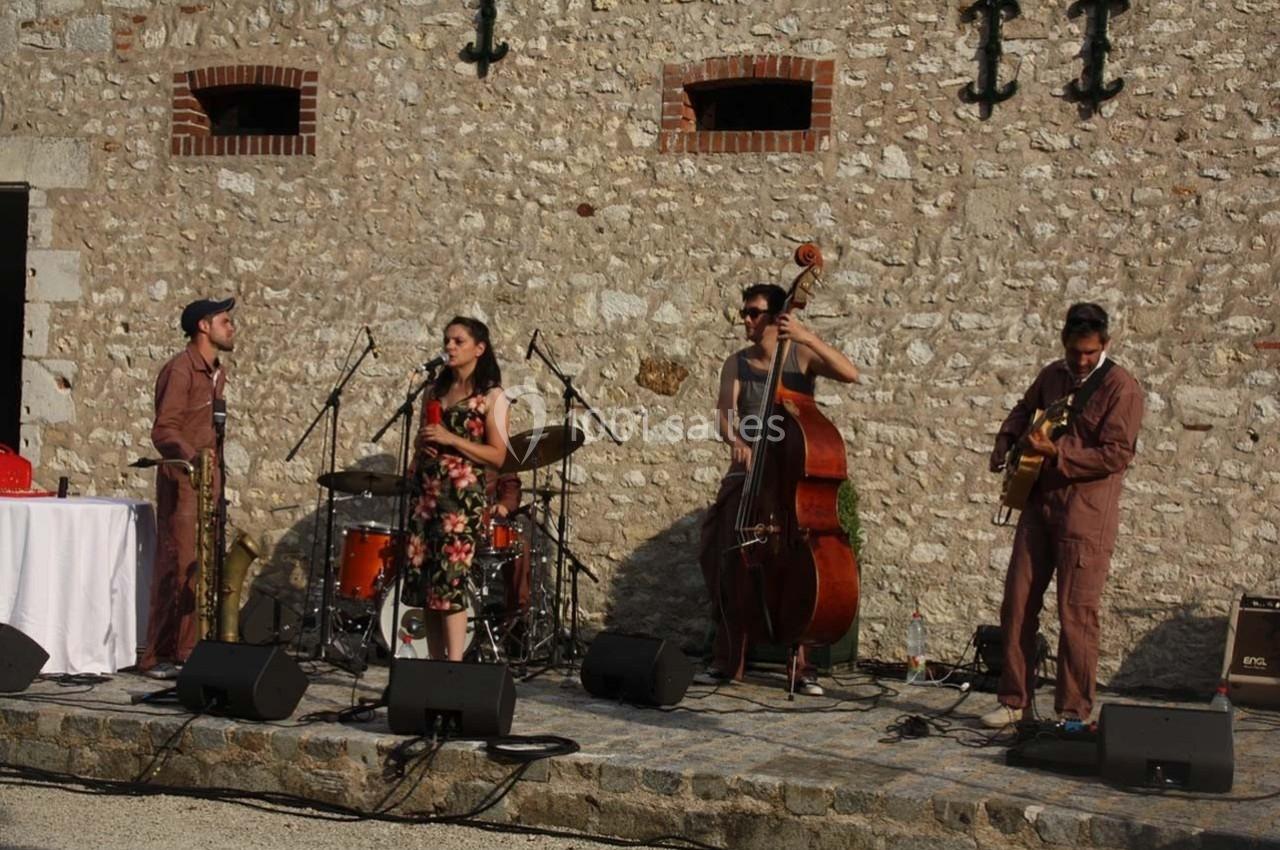 Un groupe de musiciens joue en plein air devant un mur de pierre, avec batterie, contrebasse, guitare et chant.