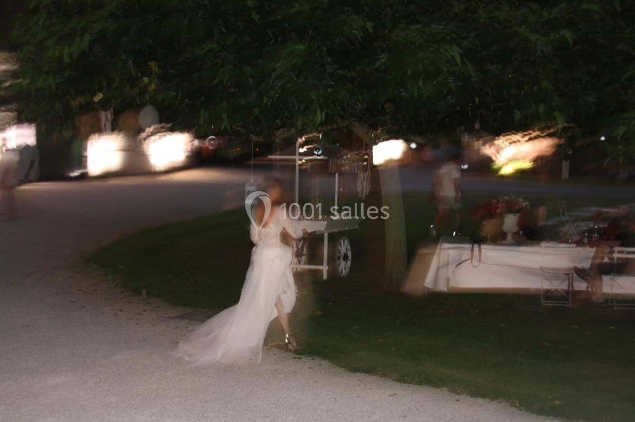 Une personne en robe blanche marche près d'un arbre dans un environnement extérieur éclairé en soirée.