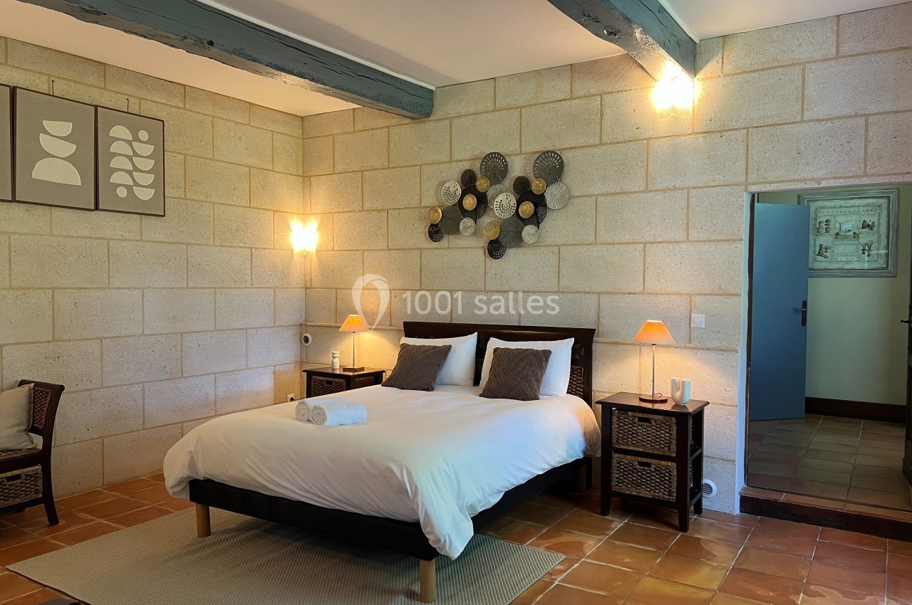 Chambre lumineuse avec lit double, murs en pierre, poutres apparentes, décoration murale et sol en terre cuite.