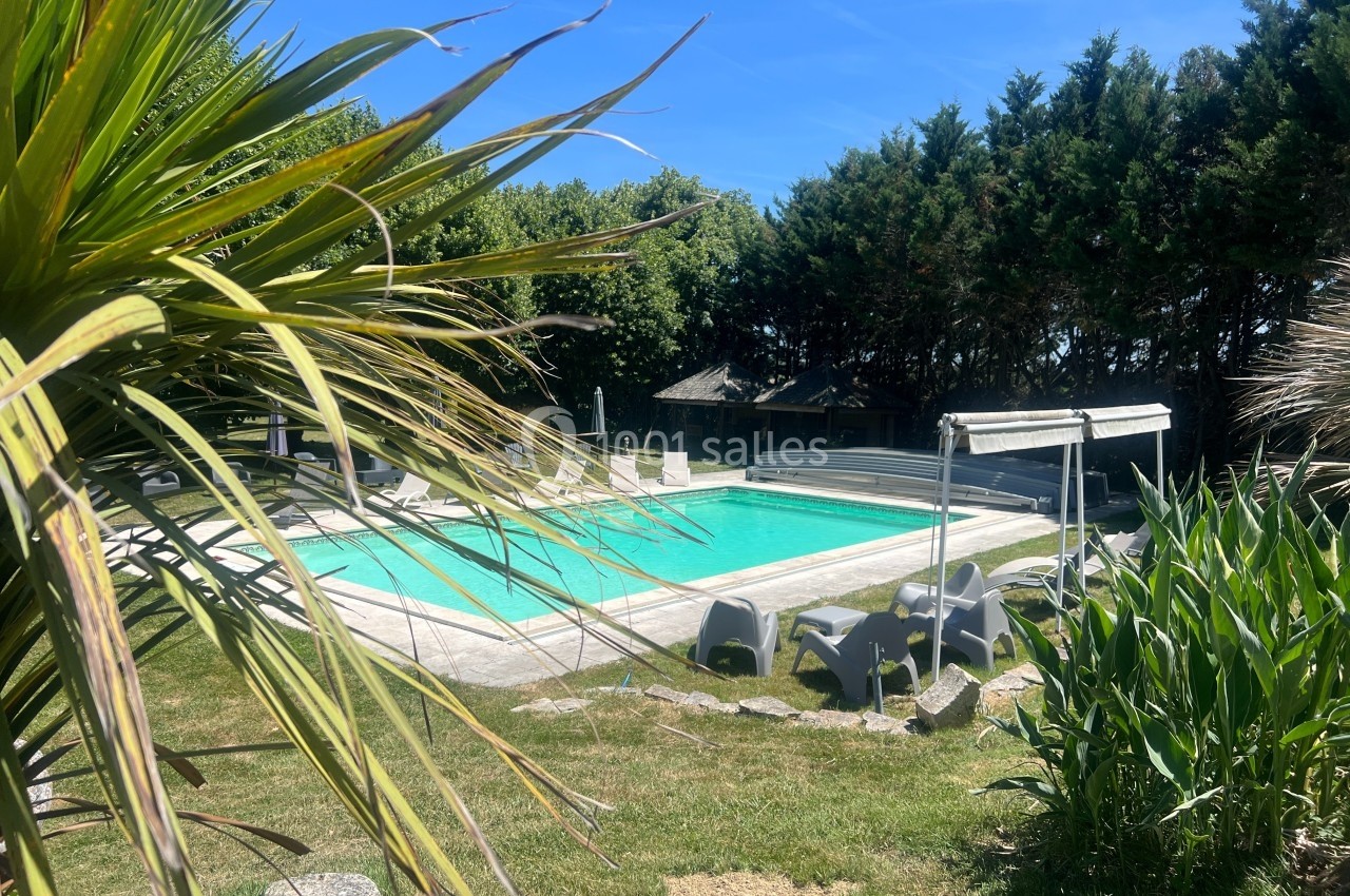 Piscine extérieure entourée de végétation, avec chaises longues et arbres en arrière-plan par une journée ensoleillée.