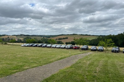 Alignement de voitures garées sur une pelouse, entourées de collines verdoyantes sous un ciel nuageux.