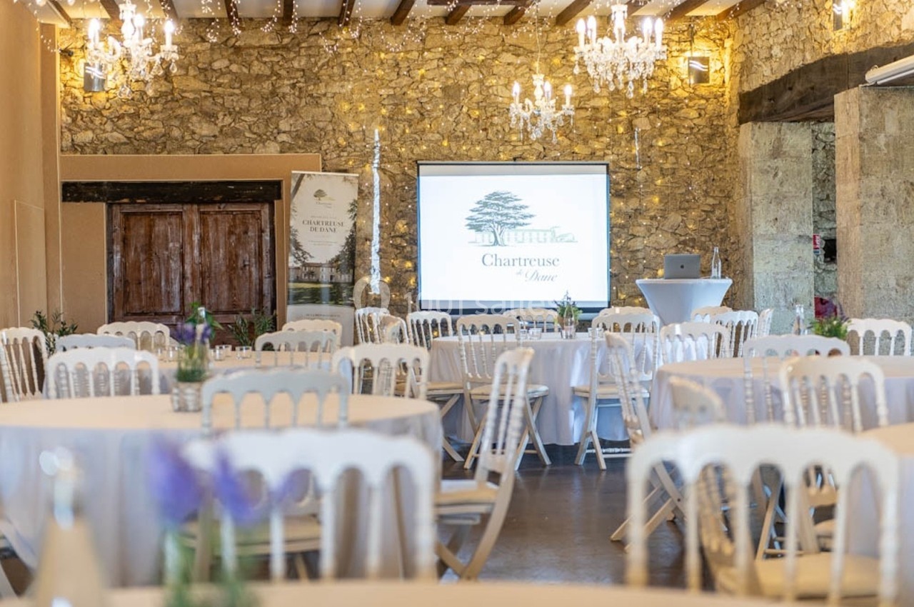 Salle de réception en pierre avec tables rondes dressées, chaises blanches et écran de projection allumé.