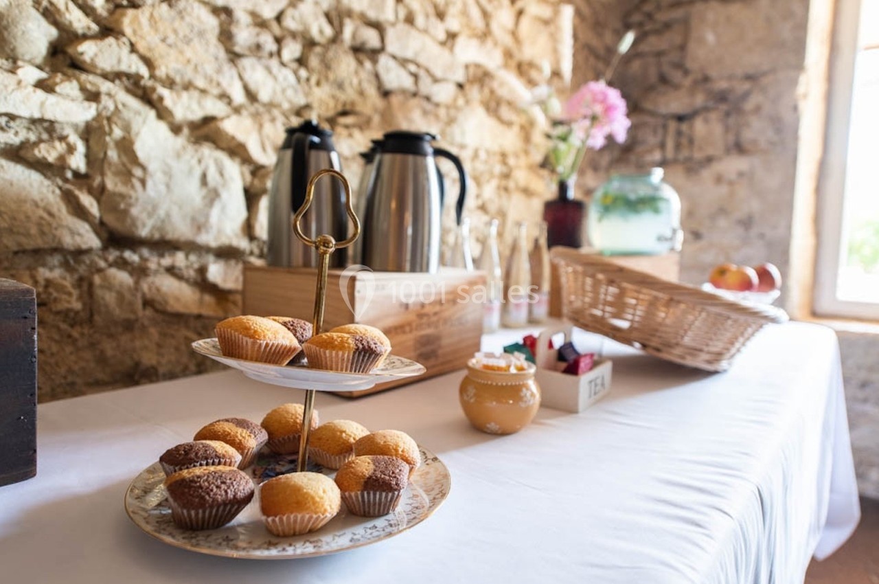 Plateau de muffins sur une table dressée avec théières, panier de fruits et décor en pierre apparente.