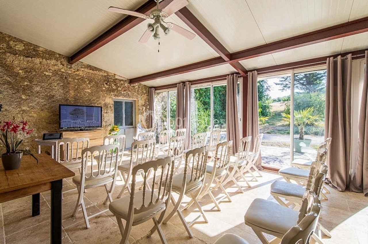 Salle lumineuse avec chaises blanches alignées, grandes baies vitrées donnant sur un jardin verdoyant.