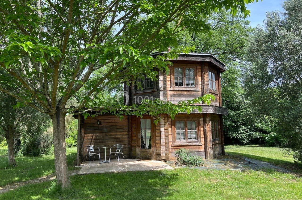 Maison en bois à deux étages entourée d'arbres, avec une terrasse équipée de chaises et d'une table.