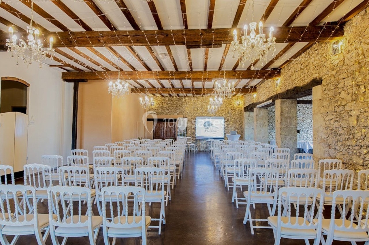 Salle de réception avec des rangées de chaises blanches alignées, murs en pierre et plafonds avec poutres apparentes.