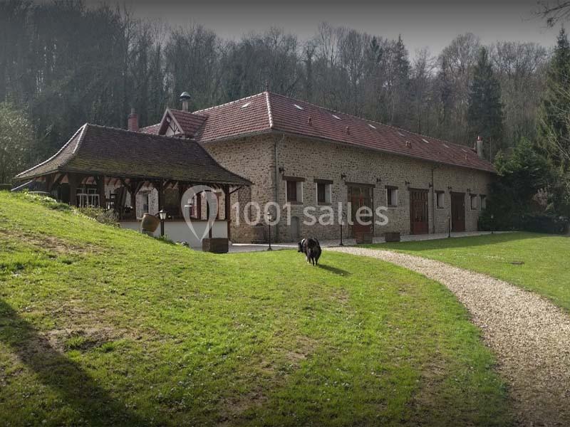 Bâtisse en pierre avec toit en tuiles, entourée de verdure, chemin en gravier et un chien sur la pelouse.