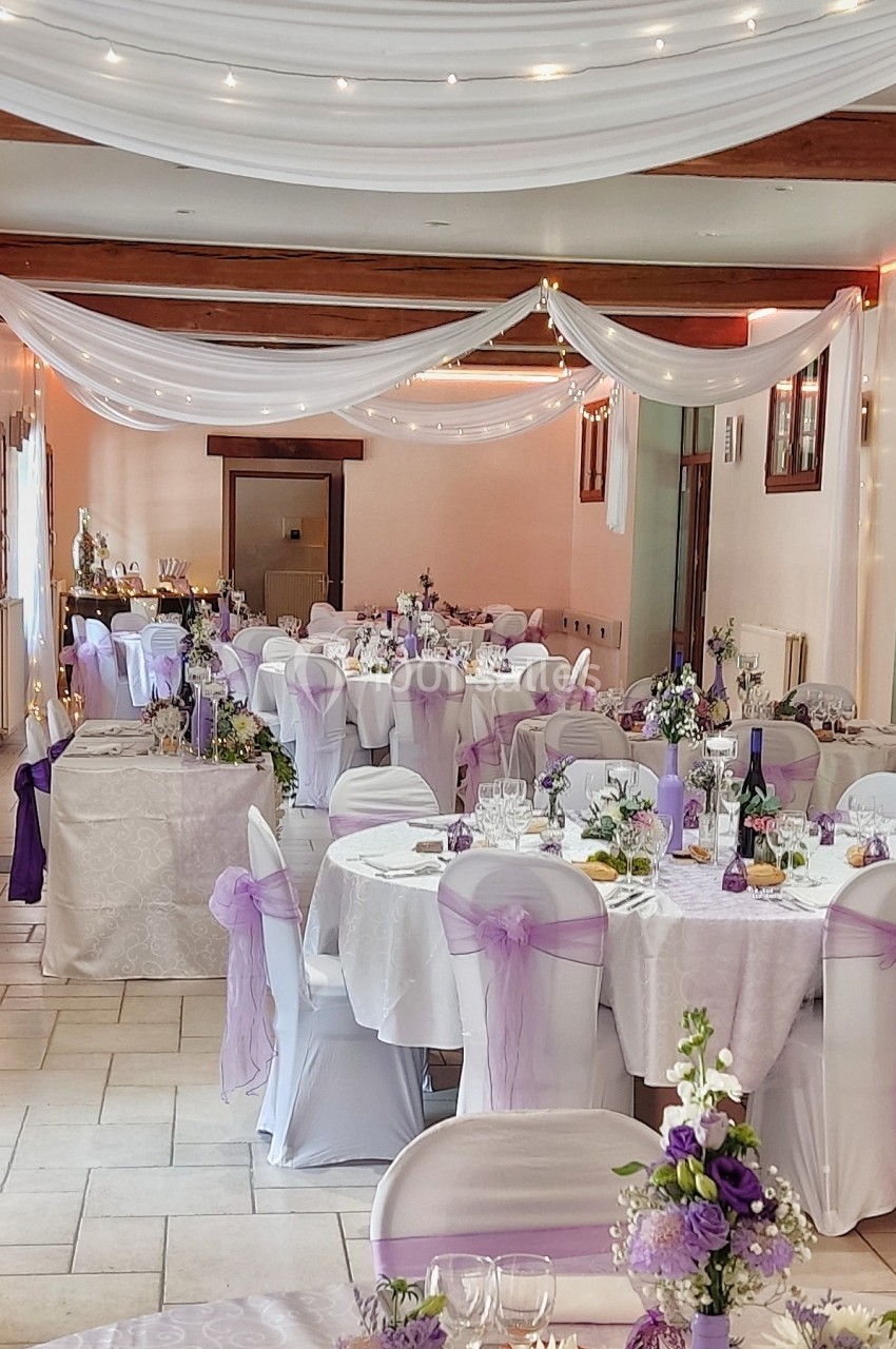 Salle de réception décorée pour un mariage avec nappes blanches, chaises ornées de rubans violets et guirlandes lumineuses.