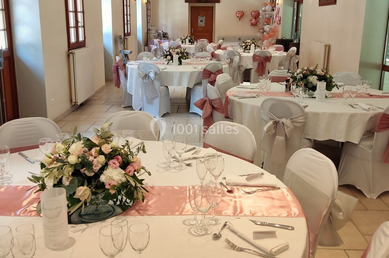 Salle de réception décorée pour un mariage, avec tables rondes dressées, nappes blanches et accents roses.